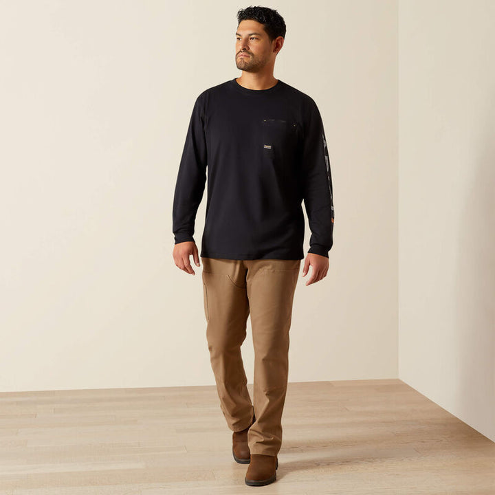 Man wearing a black long-sleeve shirt and brown pants standing in a minimalistic room.