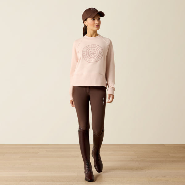 Woman wearing a pink sweatshirt, brown riding pants, and boots on a plain background