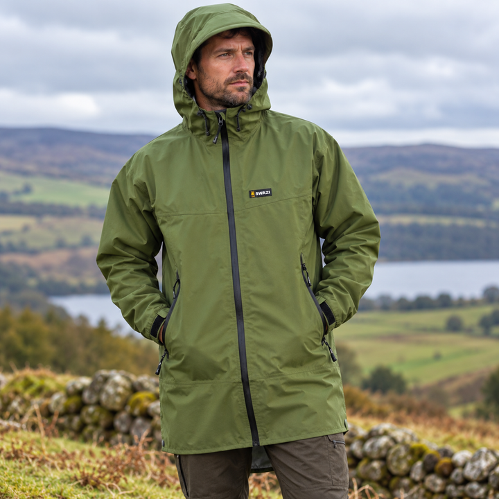 Man wearing the Swazi Men's Sentinel Ultralite Waterproof Jacket in Olive, featuring an adjustable hood, full-length zip and zipped pockets, outdoors in the countryside.