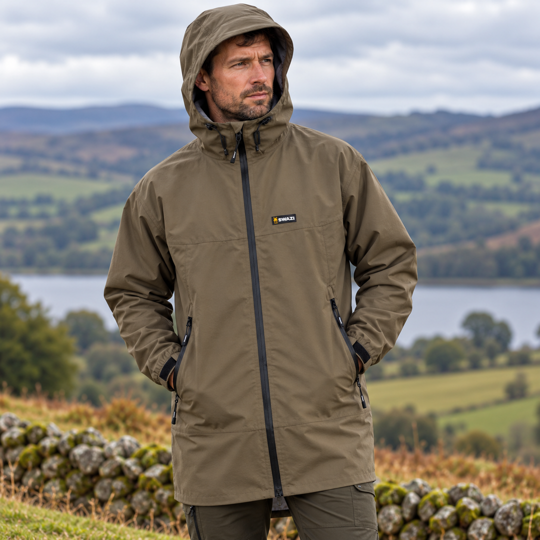 Man wearing Swazi Men's Sentinel Ultralite Waterproof Jacket in Stone, featuring an adjustable hood, full-length zip, zipped hand pockets and Swazi chest logo, outdoors in the countryside.