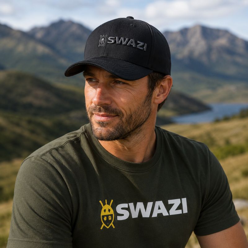 Man wearing a black cap and green t-shirt with 'SWAZI' branding in a mountainous landscape