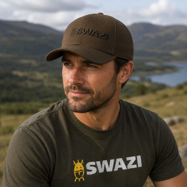 Man wearing a brown cap and black t-shirt with 'SWAZI' branding, standing in a scenic outdoor setting.