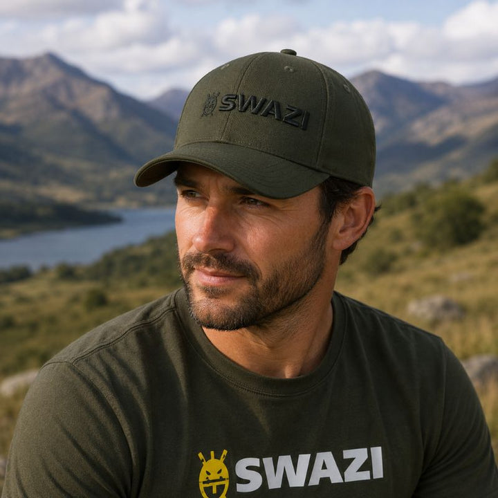 Man wearing a green 'SWAZI' cap and shirt in a mountainous landscape